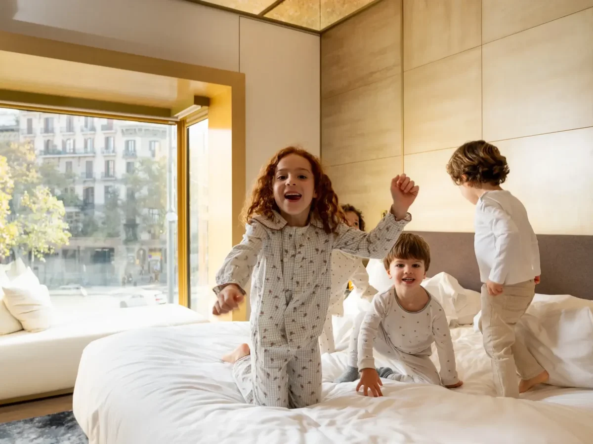 Drei Kinder in Schlafanzügen spielen und hüpfen auf einem Bett in der hell erleuchteten Almanac Family Suite, in der das Sonnenlicht durch ein großes Fenster fällt.