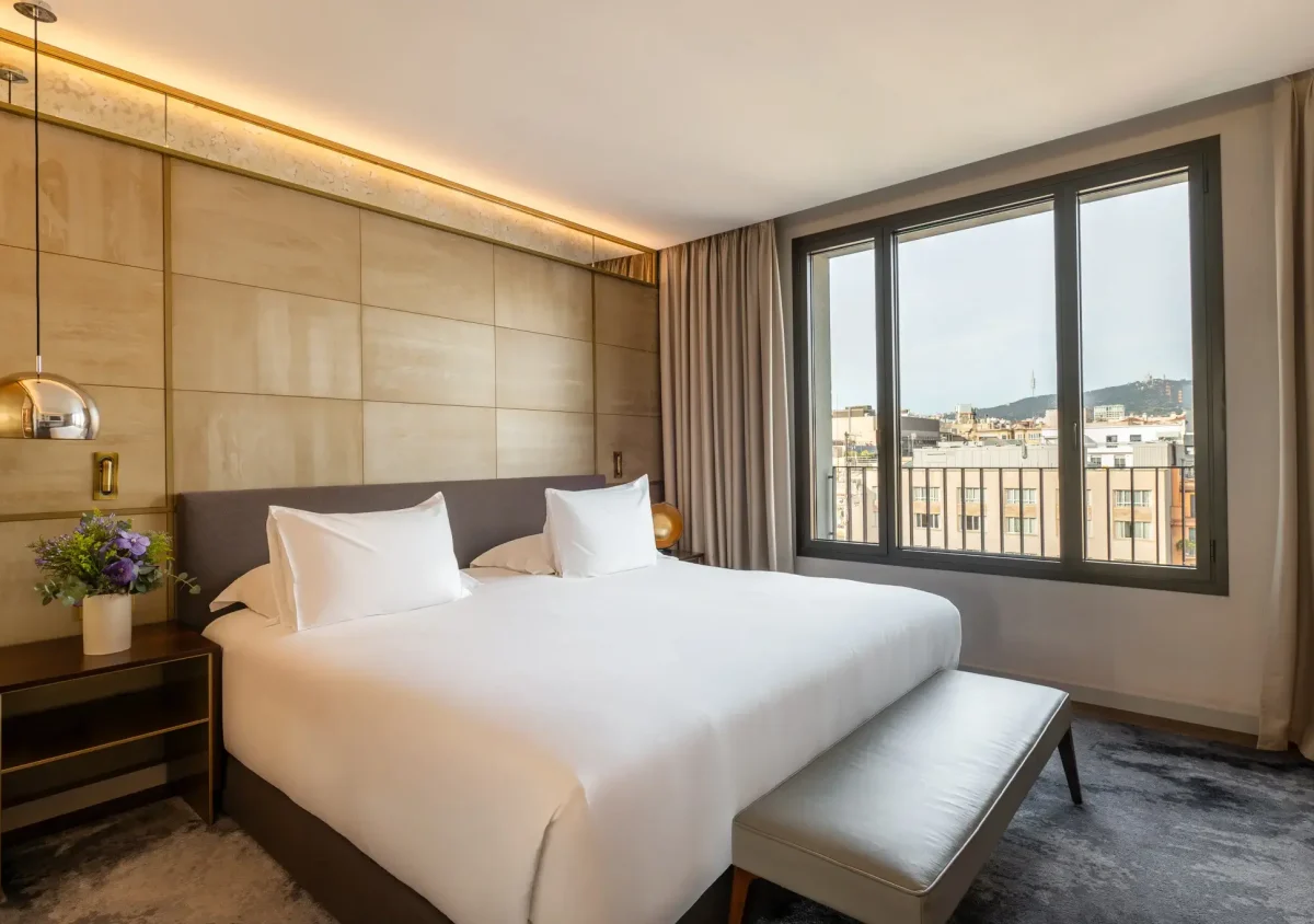 A modern hotel room with a large bed, white linens, bedside tables, a bench at the foot of the bed, and a window overlooking city buildings—a perfect choice for families seeking Family Connecting Rooms.