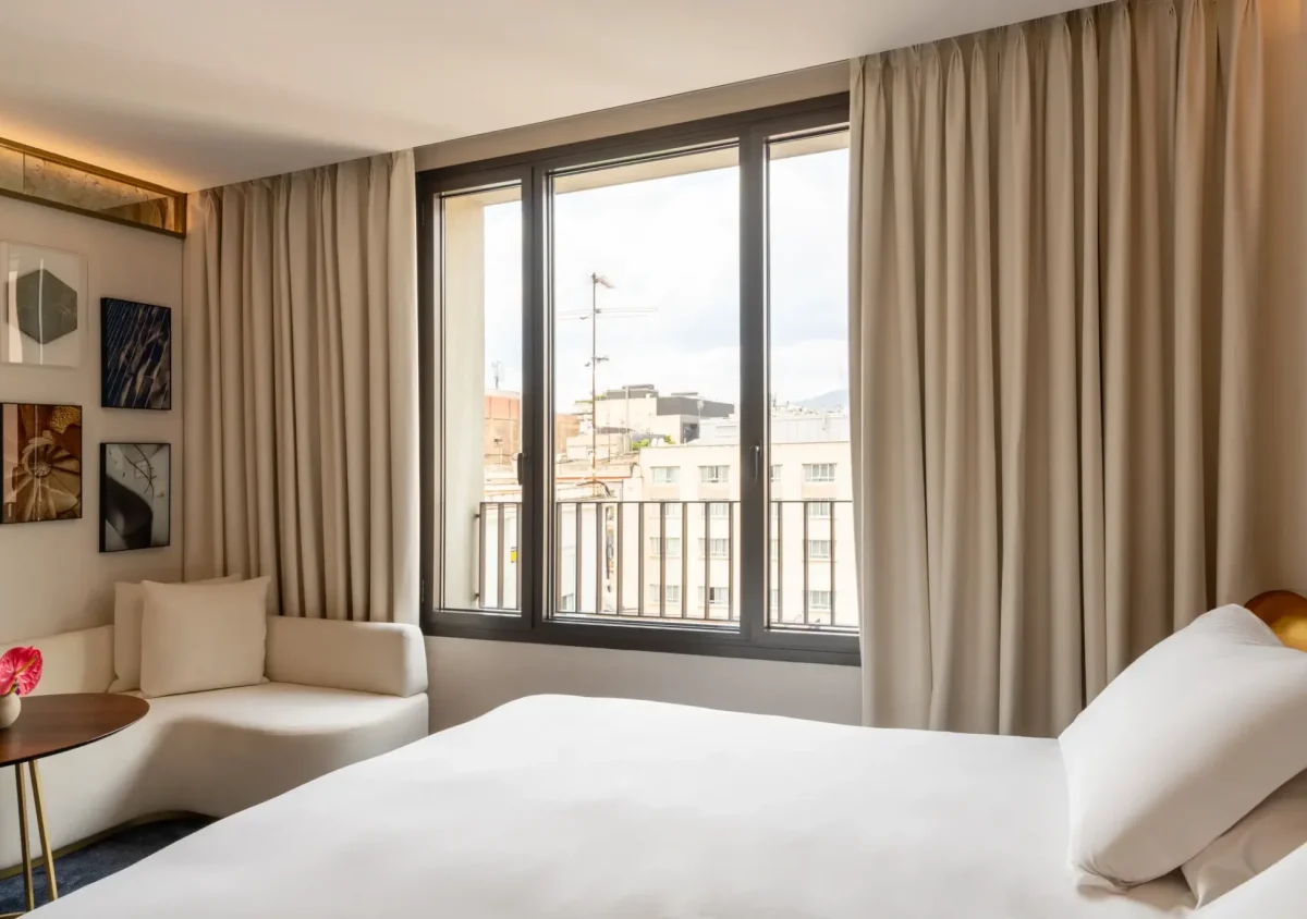 A modern hotel room with a large bed, a white corner sofa, and floor-to-ceiling windows overlooking city buildings. Family Connecting Rooms are available, making it ideal for families seeking comfort and convenience.