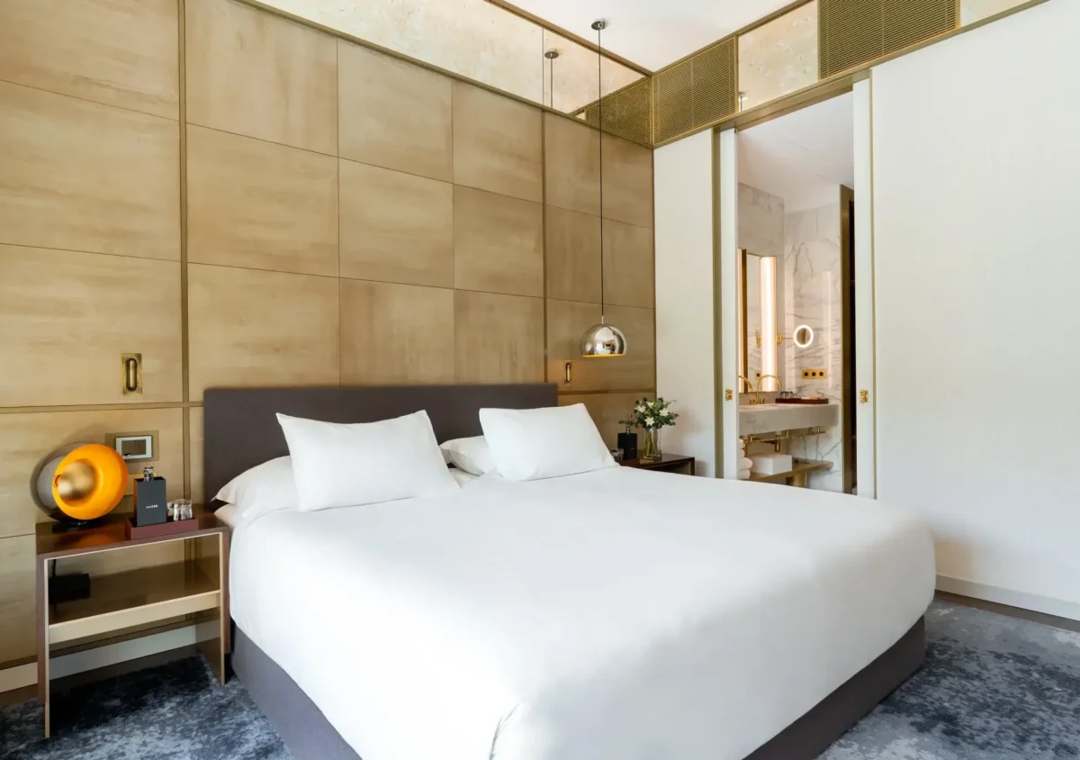 Modern bedroom with a large bed, white bedding, two bedside tables, contemporary lamps, and an open doorway leading to a bathroom—perfect for families seeking connecting rooms.