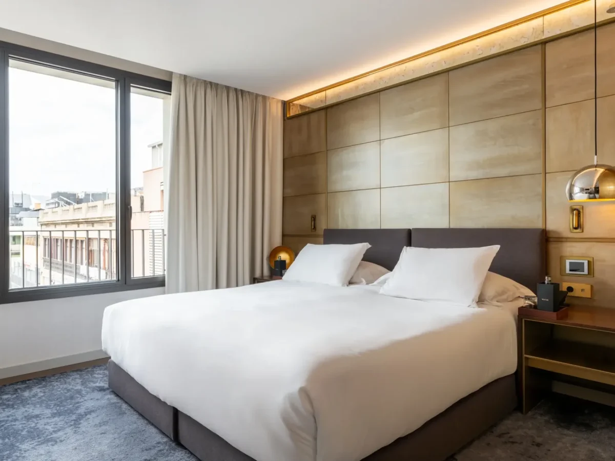 Una moderna habitación familiar comunicada con una gran cama con ropa de cama blanca, dos mesillas de noche con lámparas, amplias ventanas y elegantes paneles de pared de color beige.
