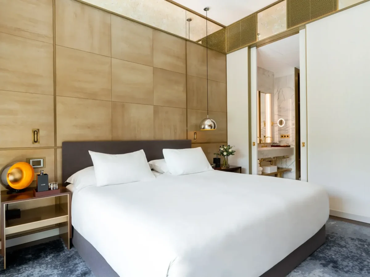 Moderne Almanac-Familiensuite mit einem großen Bett mit weißer Bettwäsche, Nachttischen, Hängeleuchten und einer offenen Tür zu einem mit Marmorwänden verzierten Badezimmer.