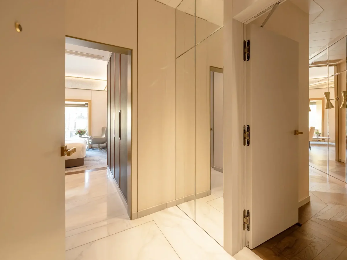 A hallway with marble floors, beige walls, mirrored panels, and open doors leading to a living area and other rooms in the Almanac Suite, featuring modern decor and abundant natural light.