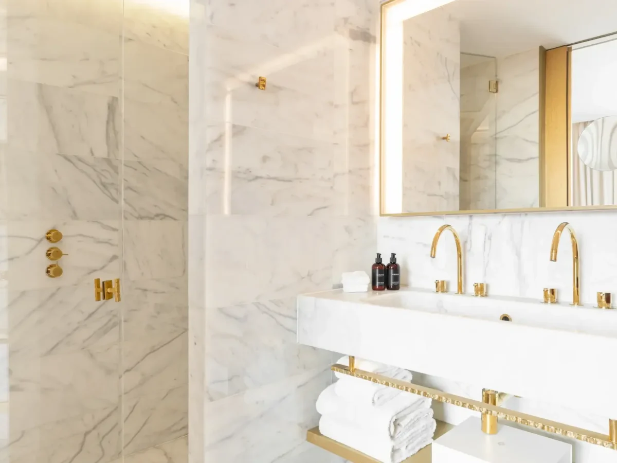Modern Gallery Room bathroom with white marble walls, gold fixtures, a double sink with two faucets, neatly folded white towels, and a glass-enclosed shower.