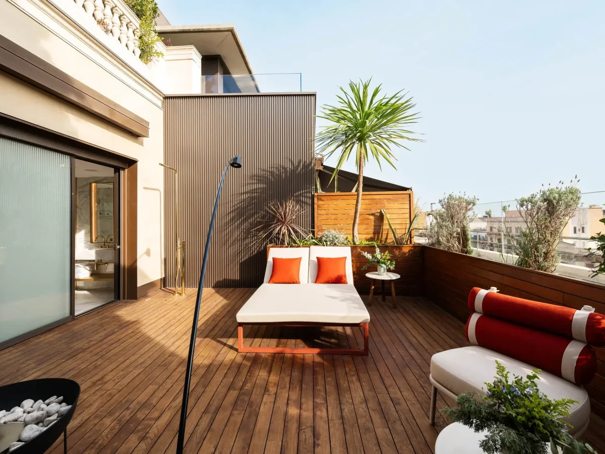 Modern rooftop terrace in the Grand Penthouse Suite with wooden flooring, a daybed with orange pillows, potted plants, outdoor chairs, and city buildings in the background under a clear sky.