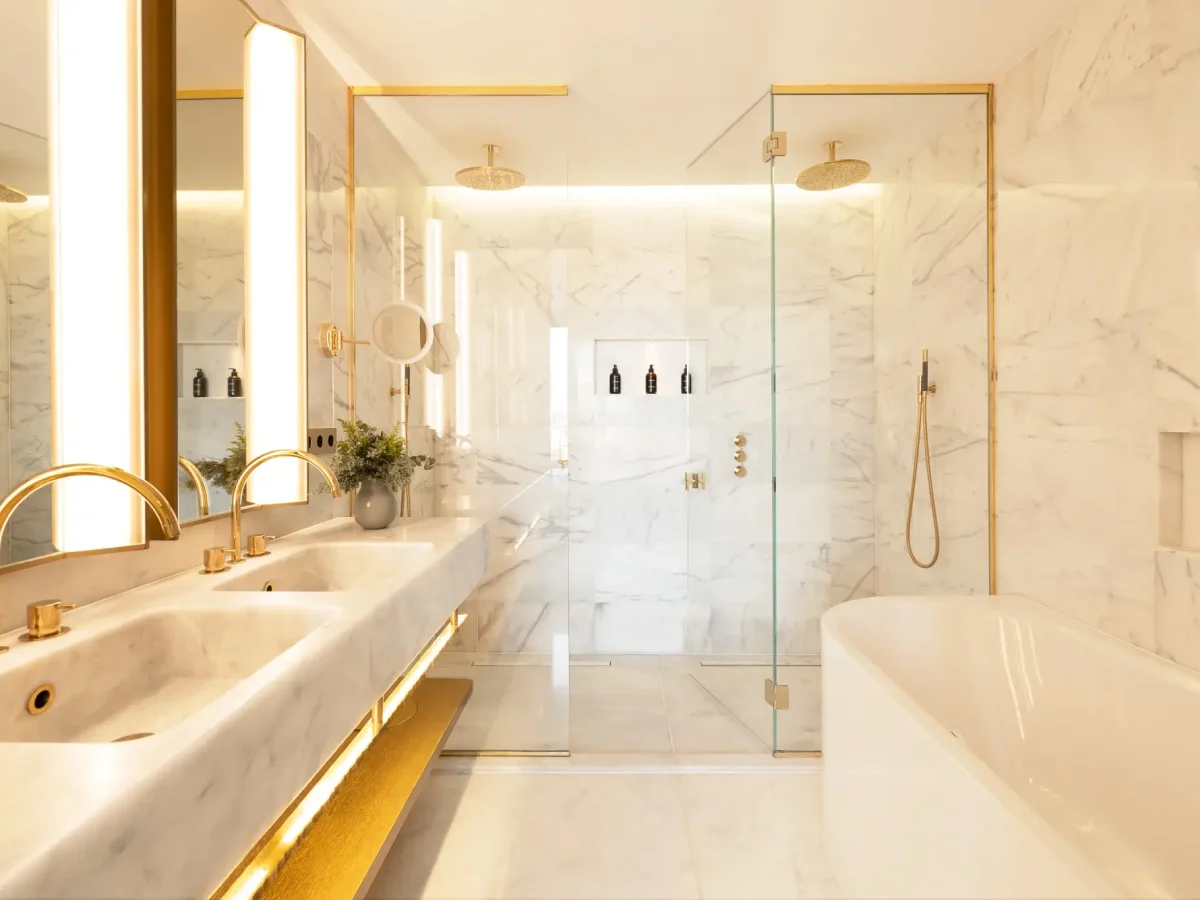 Modern bathroom in the Grand Penthouse Suite features marble walls, double sinks, gold fixtures, glass-enclosed shower, and a white bathtub. Shelves hold black toiletries, with bright, even lighting throughout.