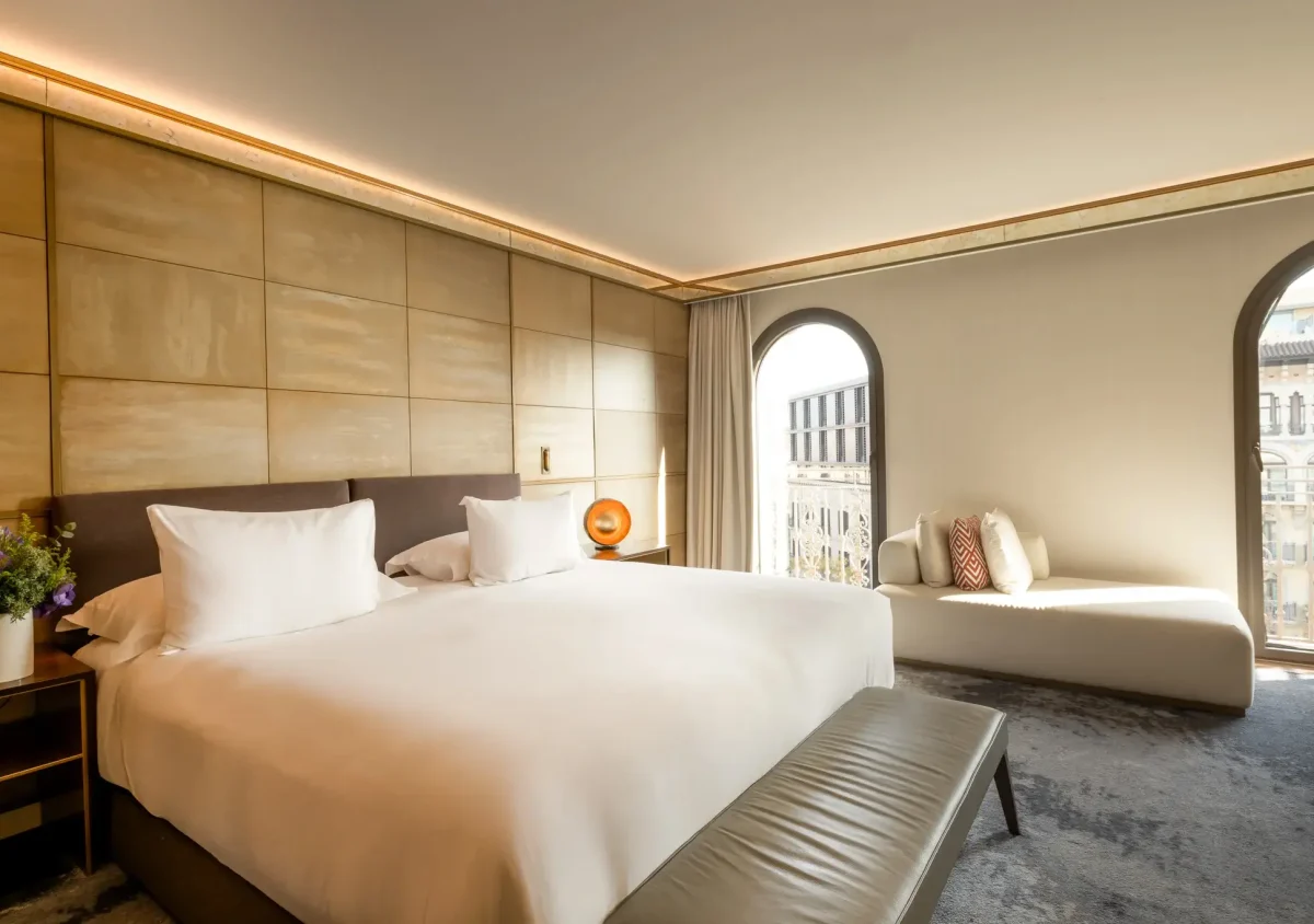 Modern Junior Suite featuring a large bed, a bench at the foot, a side table with flowers, and a cushioned lounge by arched windows bathed in soft natural light.