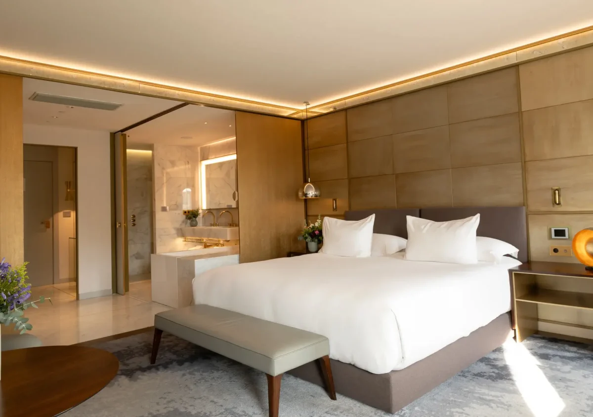 Modern Junior Suite with a large bed, white bedding, leather bench, bedside tables, fresh flowers, and a view into a bathroom featuring marble finishes and a luxurious bathtub.