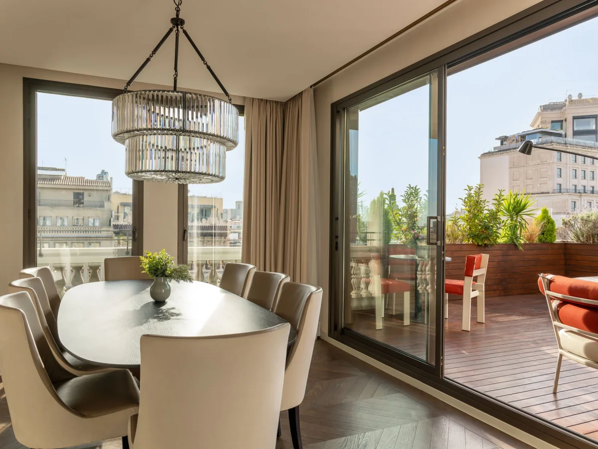 Moderno comedor en el ático con mesa ovalada y ocho sillas, gran lámpara de araña, ventanales que van del suelo al techo y un balcón con asientos al aire libre e impresionantes vistas de la ciudad.