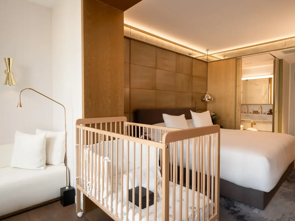 Una habitación de hotel con una cama doble, una cuna de madera, un sofá blanco y vistas a la zona del cuarto de baño con lavabo y espejo.