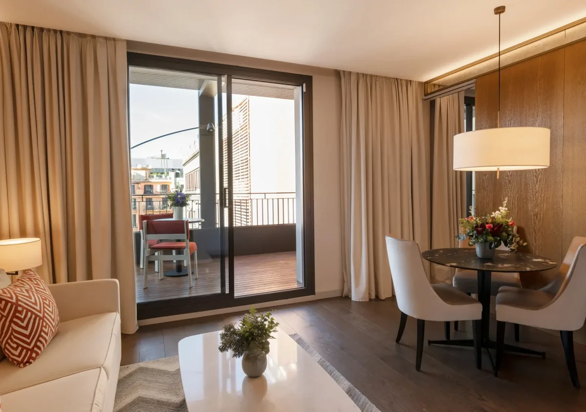 A modern Terrace Suite living room with a sofa, coffee table, small dining area, and large sliding glass doors leading to a balcony with outdoor seating.