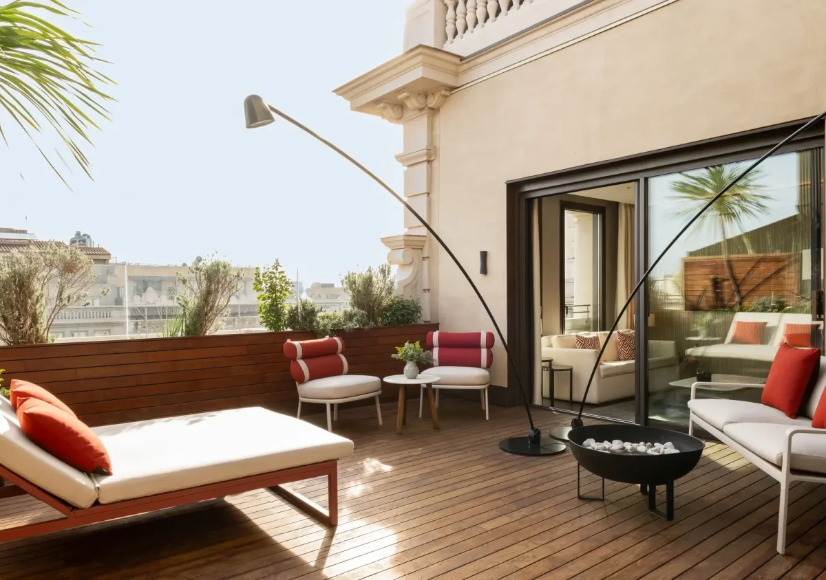 Experience the Terrace Suite: a modern terrace with wooden flooring, lounge chairs, a fire pit, red and white seating, lush green plants, and large glass doors inviting you inside.