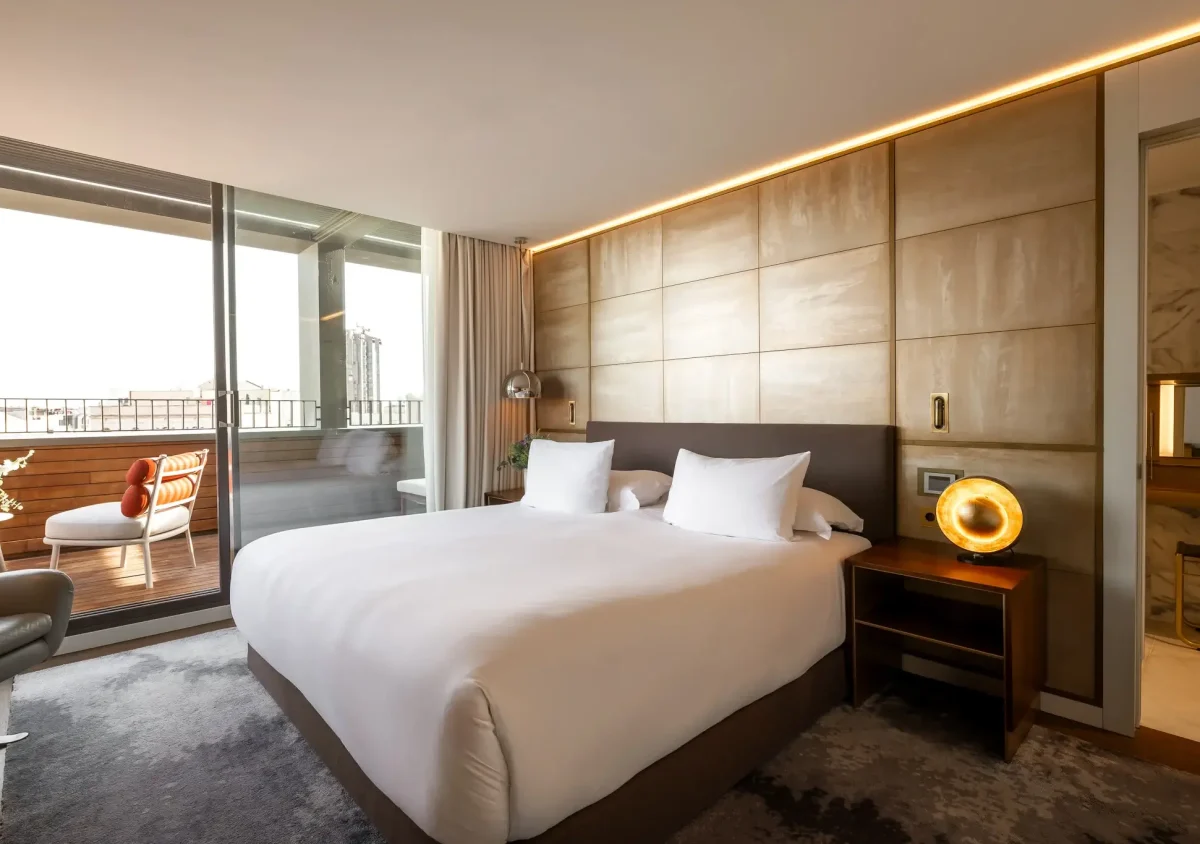 Modern Terrace Suite with a large bed, nightstand, lamp, and balcony seating area visible through glass doors.