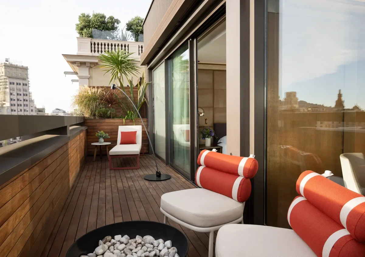 A modern Terrace Suite balcony with wooden flooring, white and orange cushioned chairs, a small lounge area, potted plants, and a stunning cityscape view in the background.