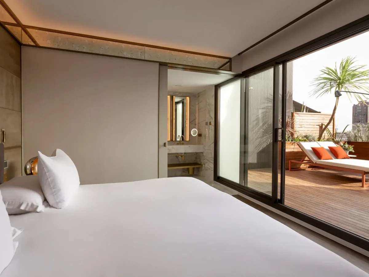 A modern Terrace Suite featuring a white bed, sliding glass doors, and a private terrace with lounge chairs and a potted palm tree.