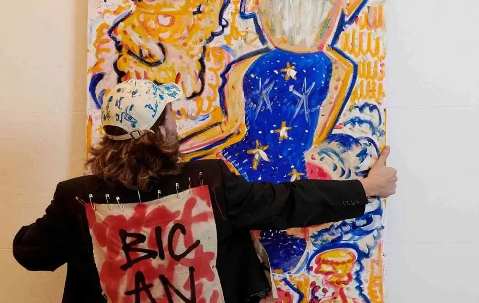 Osoba v čepici a bundě s nápisem "BIC AN" na zádech drží u zdi barevný abstraktní obraz, který evokuje živého ducha SIGNATURE TOURS.