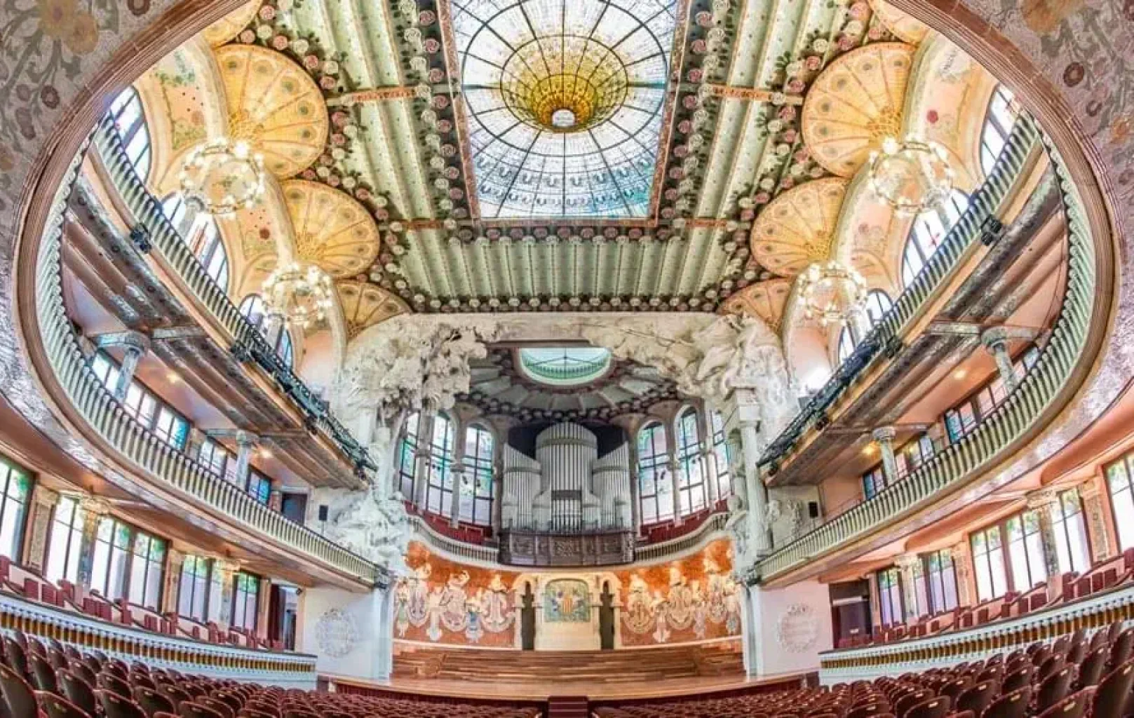 Ornamentado interior de sala de conciertos de estilo modernista catalán, con techo decorativo, cúpula de vidrieras, balcones, lámparas de araña y un órgano en el escenario frente a filas de asientos rojos vacíos: el escenario perfecto para una invitación privada.