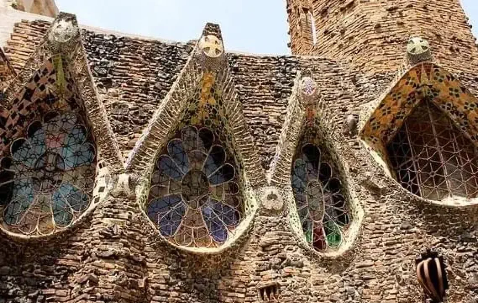 Detalle de una pared de ladrillo con cuatro vidrieras altas y arqueadas con detalles de mosaico, que muestran una textura irregular y rústica inspirada en el Modernismo Catalán.