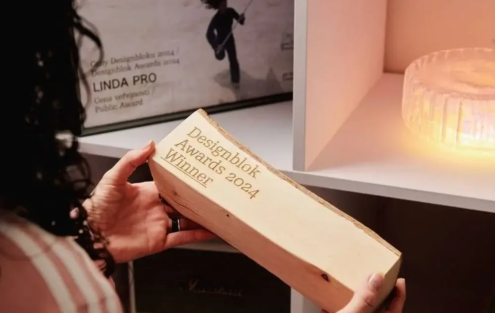 Osoba drží dřevěný blok s vyrytým nápisem "Designblok Awards 2024 Winner" v regálu plném designových předmětů, který vystihuje kreativního ducha Lindina konceptuálního obchodu.