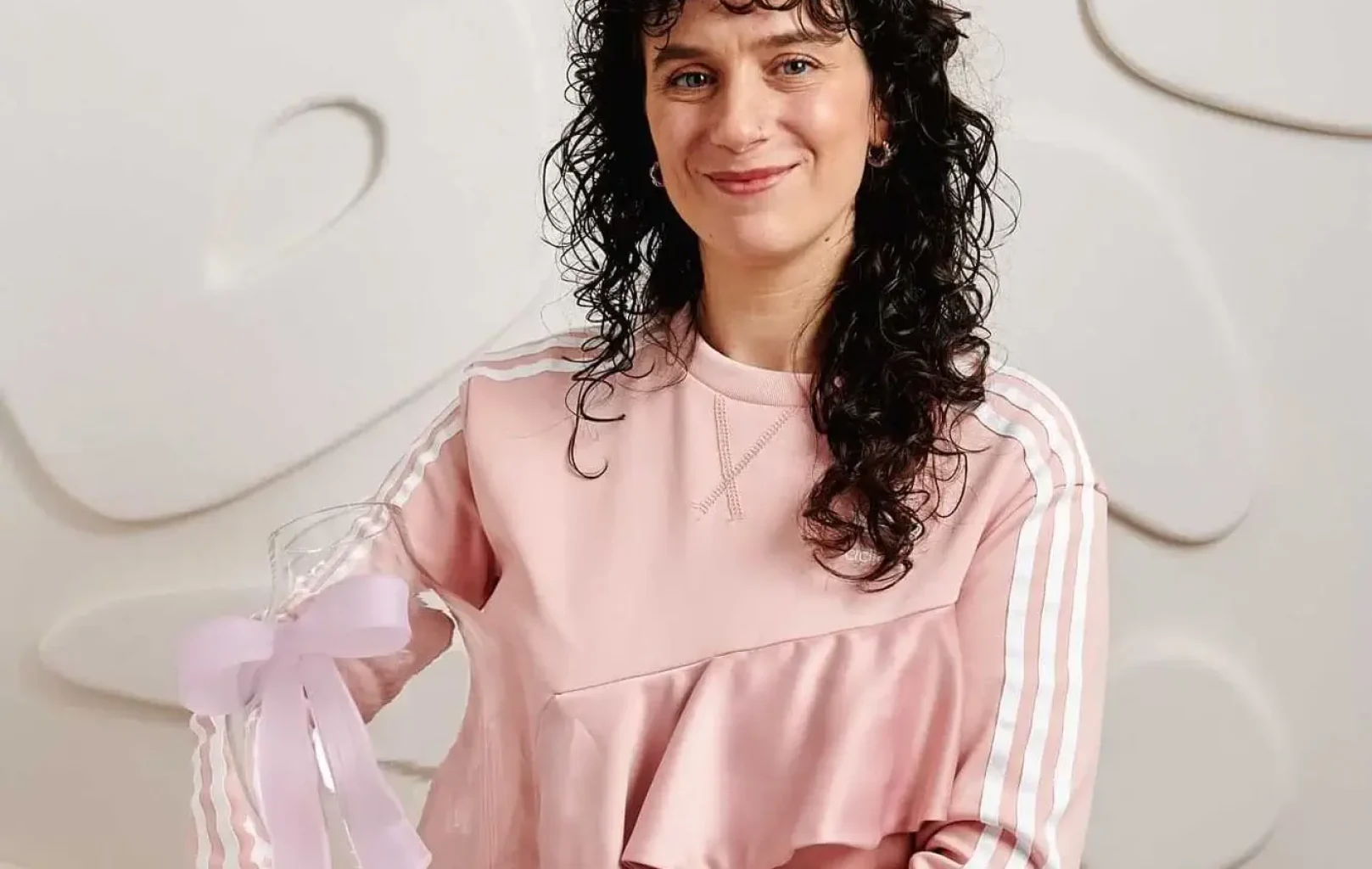 Žena s kudrnatými tmavými vlasy v růžovém oblečení drží průhledné ocenění s levandulovou stuhou a stojí před bílou texturovanou stěnou, která oslavuje úspěchy designérky Lindy Pro na akci concept store.