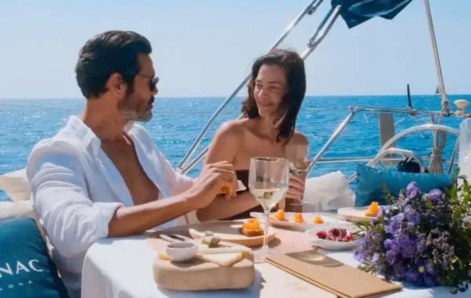 A man and woman sit at a table on a boat, enjoying drinks and snacks with the ocean in the background, indulging in a luxurious Suite Experience.