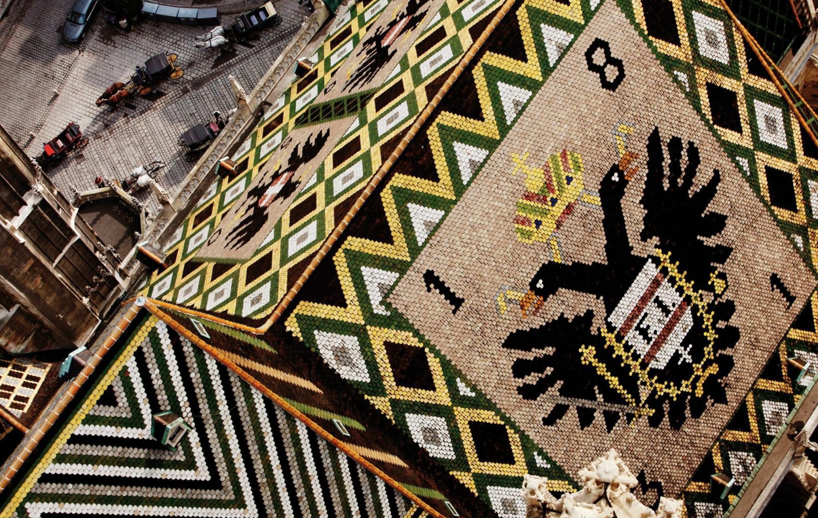 Nahaufnahme eines farbenfrohen Ziegeldachs mit einem schwarzen Doppeladler und geometrischen Mustern, das einen Blick auf den darunter liegenden Stadtplatz bietet.