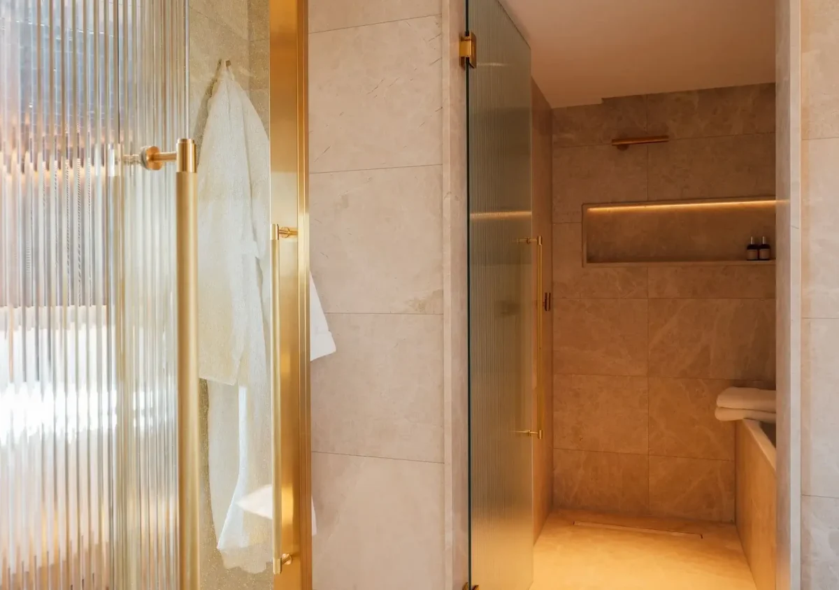 Modern bathroom in the Almanac Park Suite with beige marble walls, glass shower door, towel hanging on a hook, and built-in shelf with toiletries.