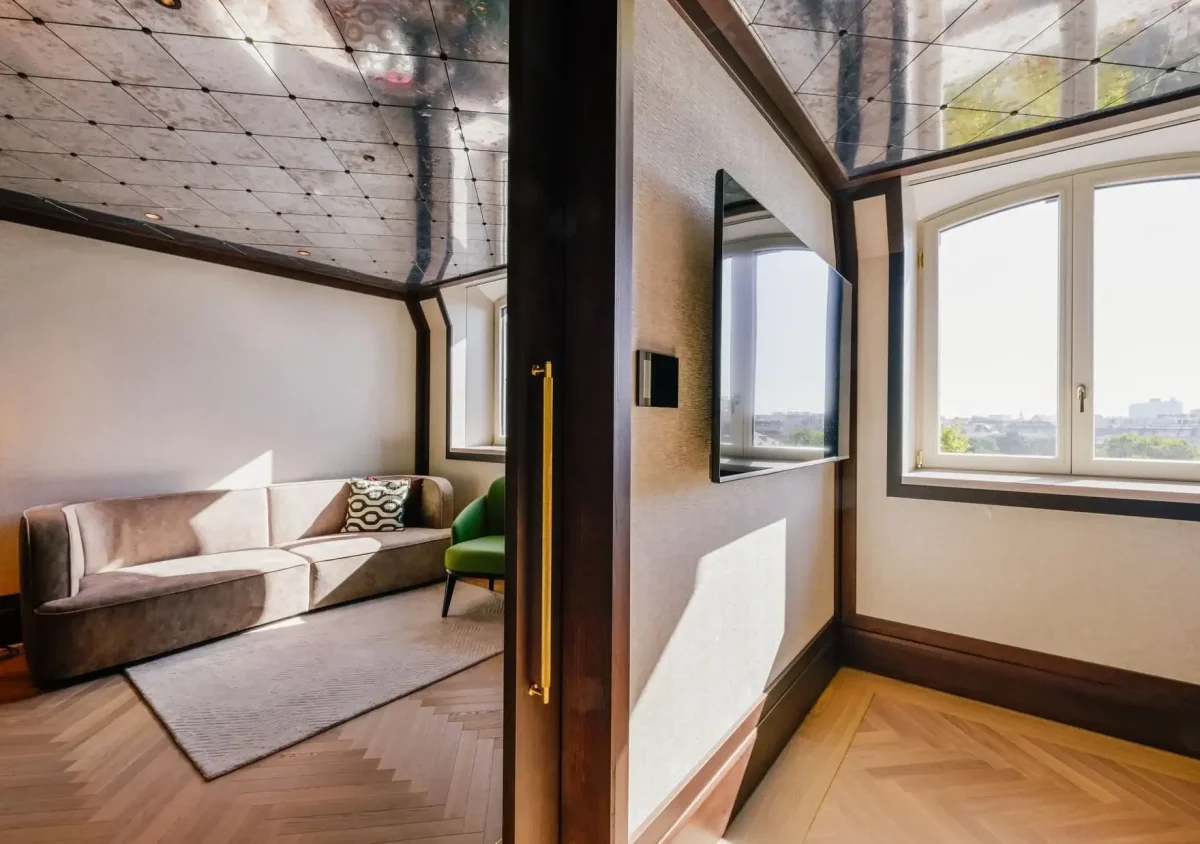 A corner of the Almanac Park Suite features a beige sofa, green chair, wall-mounted TV, large window, and a reflective ceiling that reveals striking structural elements.