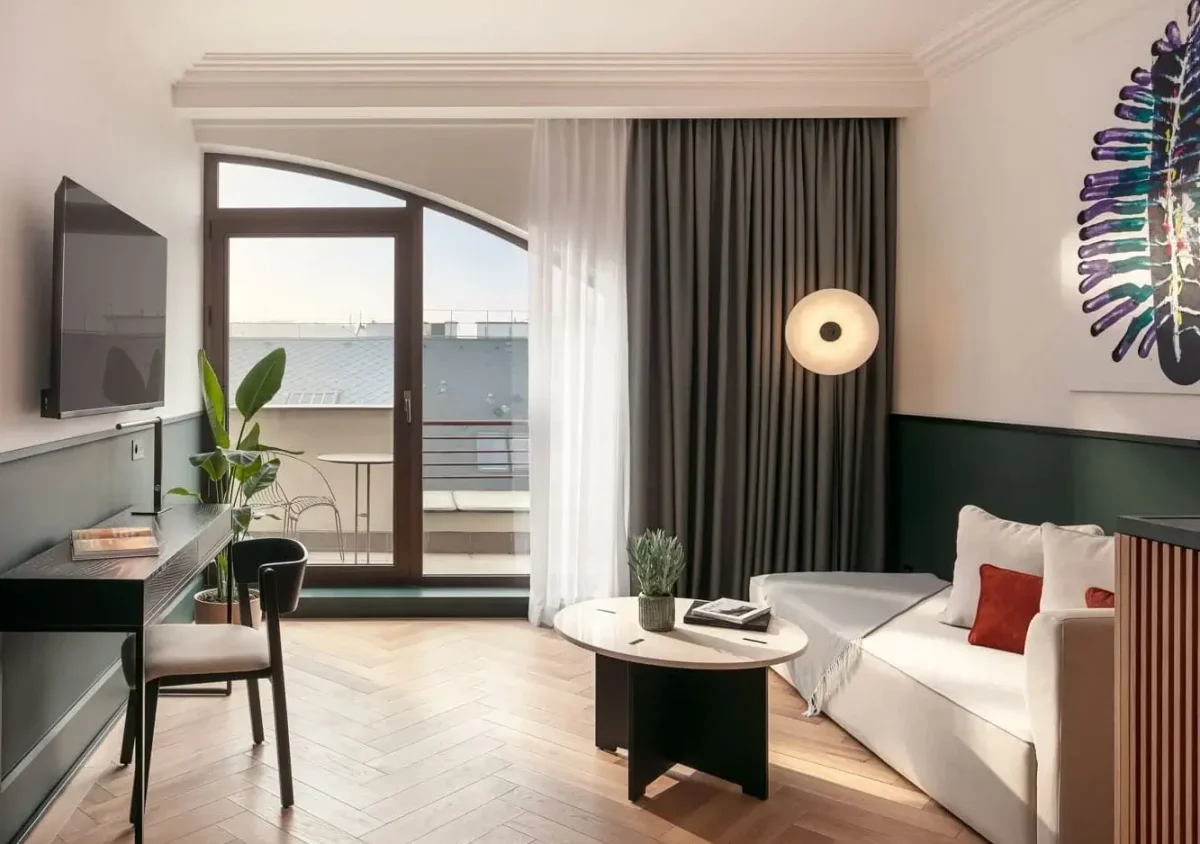 Modern living room in this Executive Suite with a white sofa, round coffee table, desk, chair, wall-mounted TV, large window with curtains leading to a balcony, potted plant, and contemporary artwork on the wall.