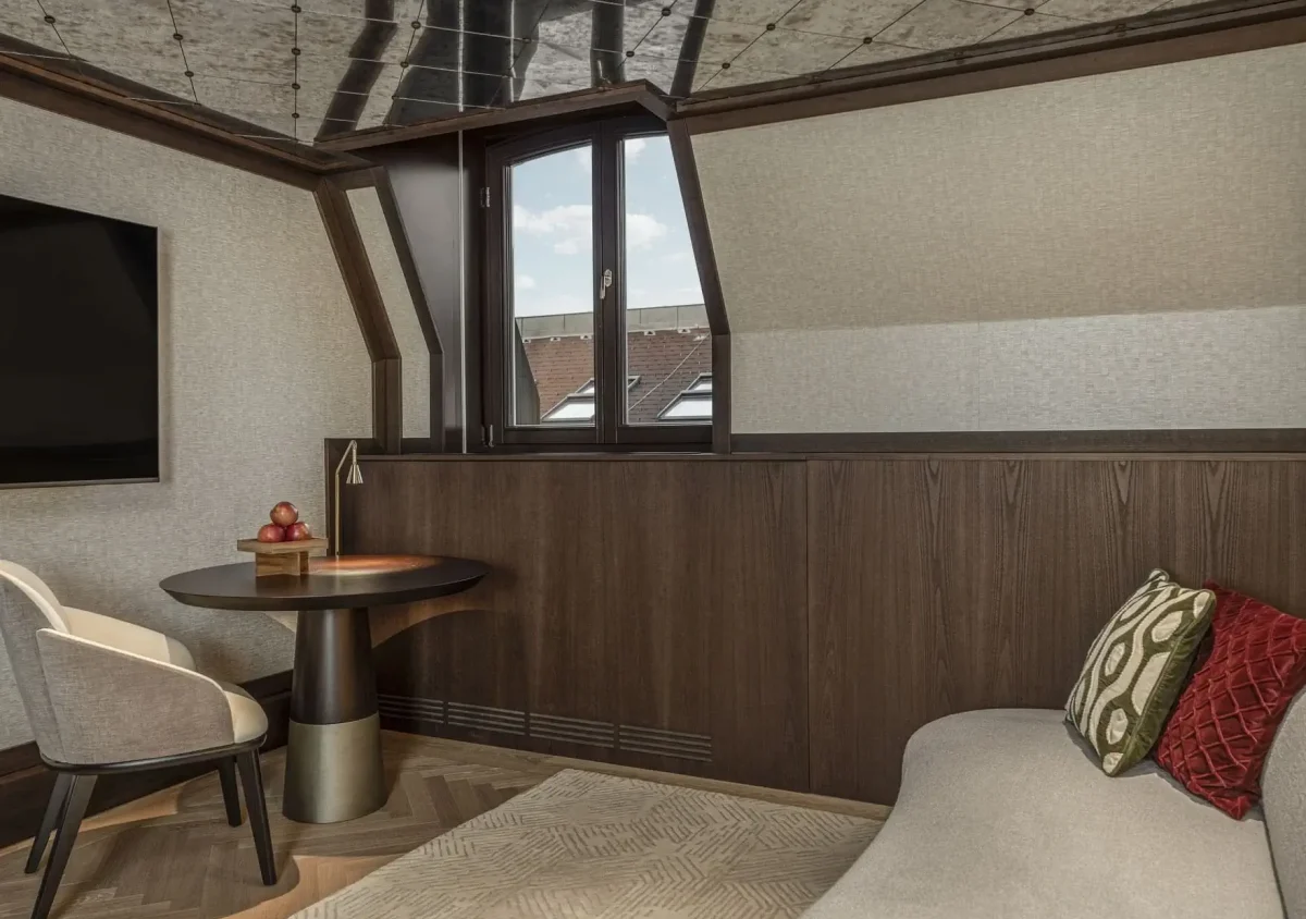 Modern attic room in the Almanac Suite with a small round table, two chairs, a flat-screen TV, a window, and a beige sofa with decorative pillows. Dark wood paneling and a patterned ceiling add character.