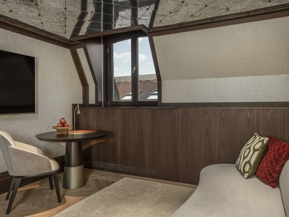 Modern attic room in the Almanac Suite with a small round table, two chairs, a flat-screen TV, a window, and a beige sofa with decorative pillows. Dark wood paneling and a patterned ceiling add character.