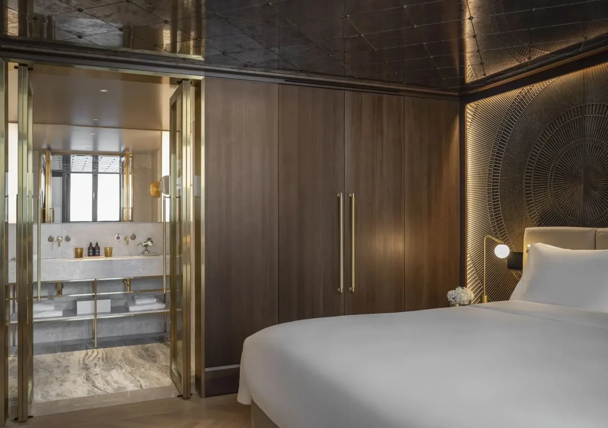 A modern Almanac Suite featuring a large bed, wood-paneled walls, decorative wall art, and a view into a marble bathroom with gold accents and glass doors.