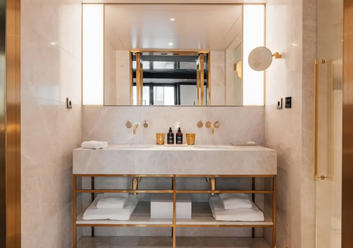 Modern bathroom in the Almanac Suite featuring a double sink vanity, gold fixtures, large mirror, under-sink shelving with towels, and elegant marble walls. Bottles and towels are thoughtfully placed on the countertop.