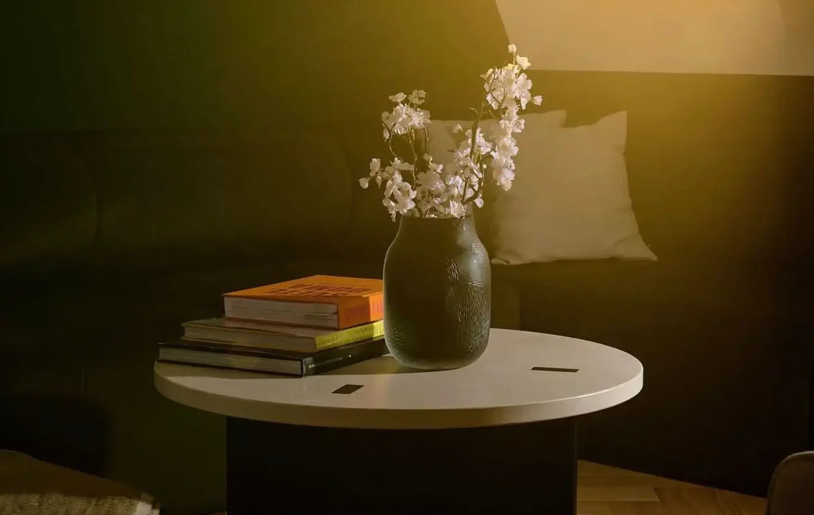 Ein runder weißer Couchtisch mit einer schwarzen Vase mit weißen Blumen und gestapelten Büchern steht im sonnendurchfluteten Wohnzimmer der PRÄSIDENTEN-SUITE, das durch ein dunkles Sofa und schräge Fenster akzentuiert wird.