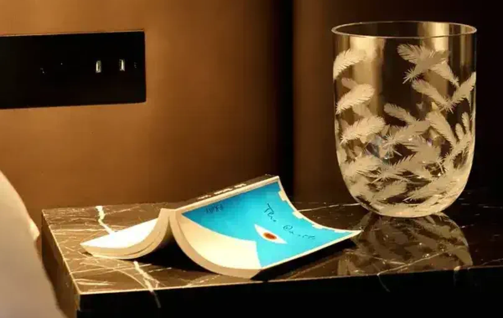 Ein aufgeschlagenes Buch liegt auf einer schwarzen Marmorplatte neben einer transparenten Vase aus Rückl-Glas, die mit einem Federmuster verziert ist und an den Luxus des Kristalltraums erinnert.