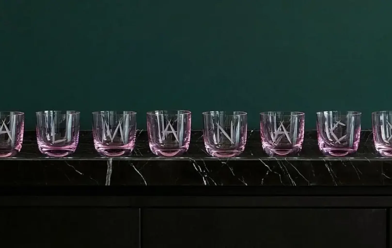Auf einer schwarzen Marmorplatte reihen sich acht RÜCKL-Klargläser mit rosafarbenen Böden aneinander, die jeweils mit einem anderen Buchstaben geätzt sind und der KRISTALLTRAUM PRÄSIDENTEN-SUITE vor dunkelgrünem Hintergrund Luxus verleihen.