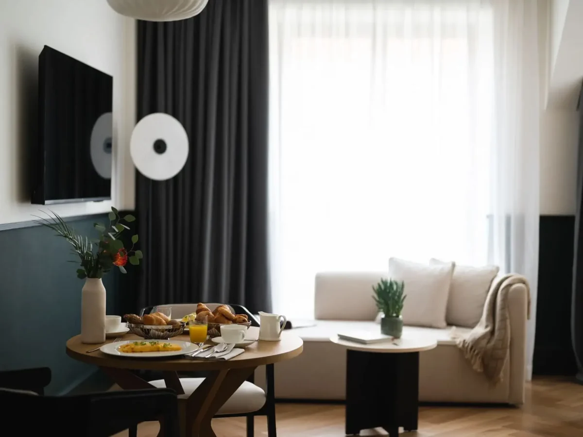 Ein modernes Wohnzimmer in der Junior Suite mit einem runden Esstisch für das Frühstück, einem weißen Sofa, einem Couchtisch mit einer Pflanze und einem großen Fenster mit durchsichtigen Vorhängen.