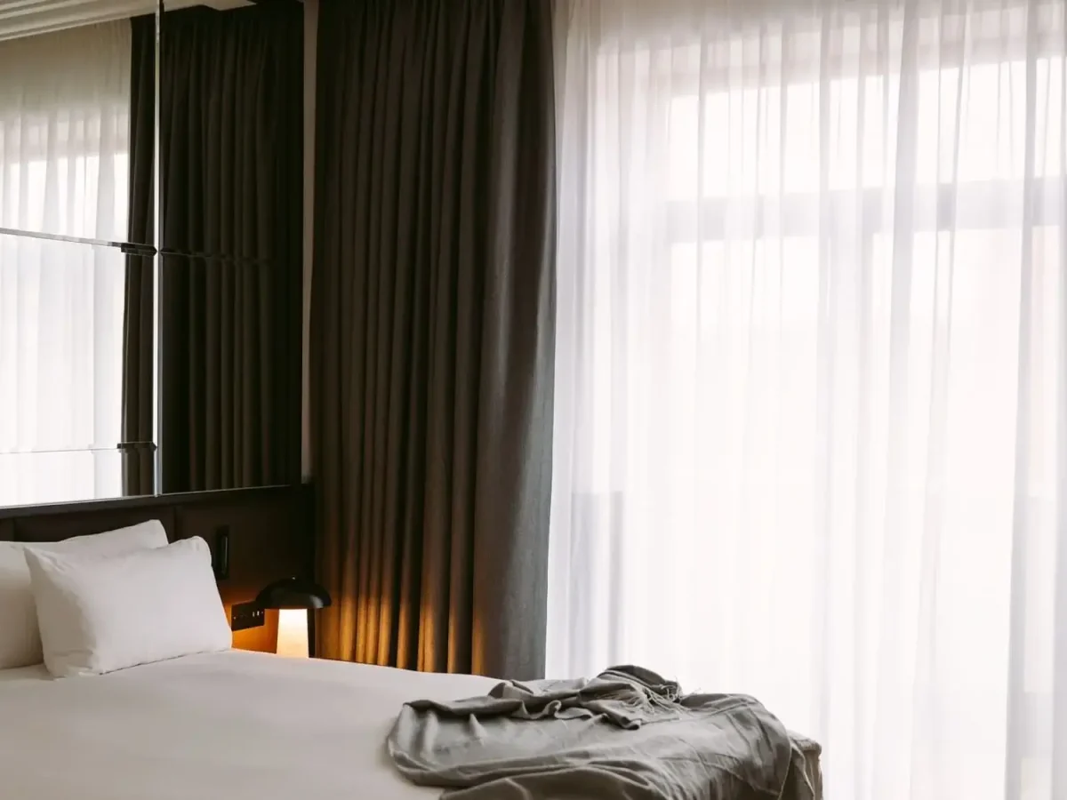 Ein ordentlich gemachtes Bett mit weißer Bettwäsche und Kissen, eine graue Decke am Fußende und große Fenster mit durchsichtigen und dunklen Vorhängen, die natürliches Licht hereinlassen, schaffen ein ruhiges Ambiente in der Executive Suite.