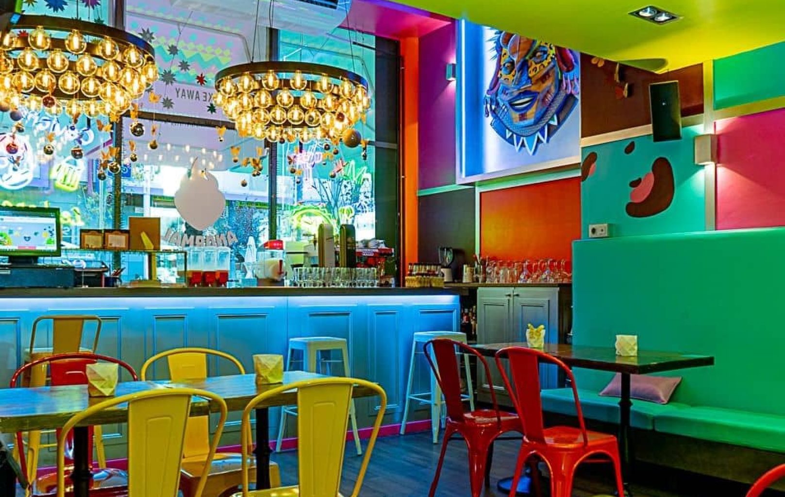 Colorful, modern cafe interior with vibrant walls, multicolored chairs, wooden tables, chandeliers, and a bar counter in the background—perfect for enjoying Anormal Food in a lively setting.