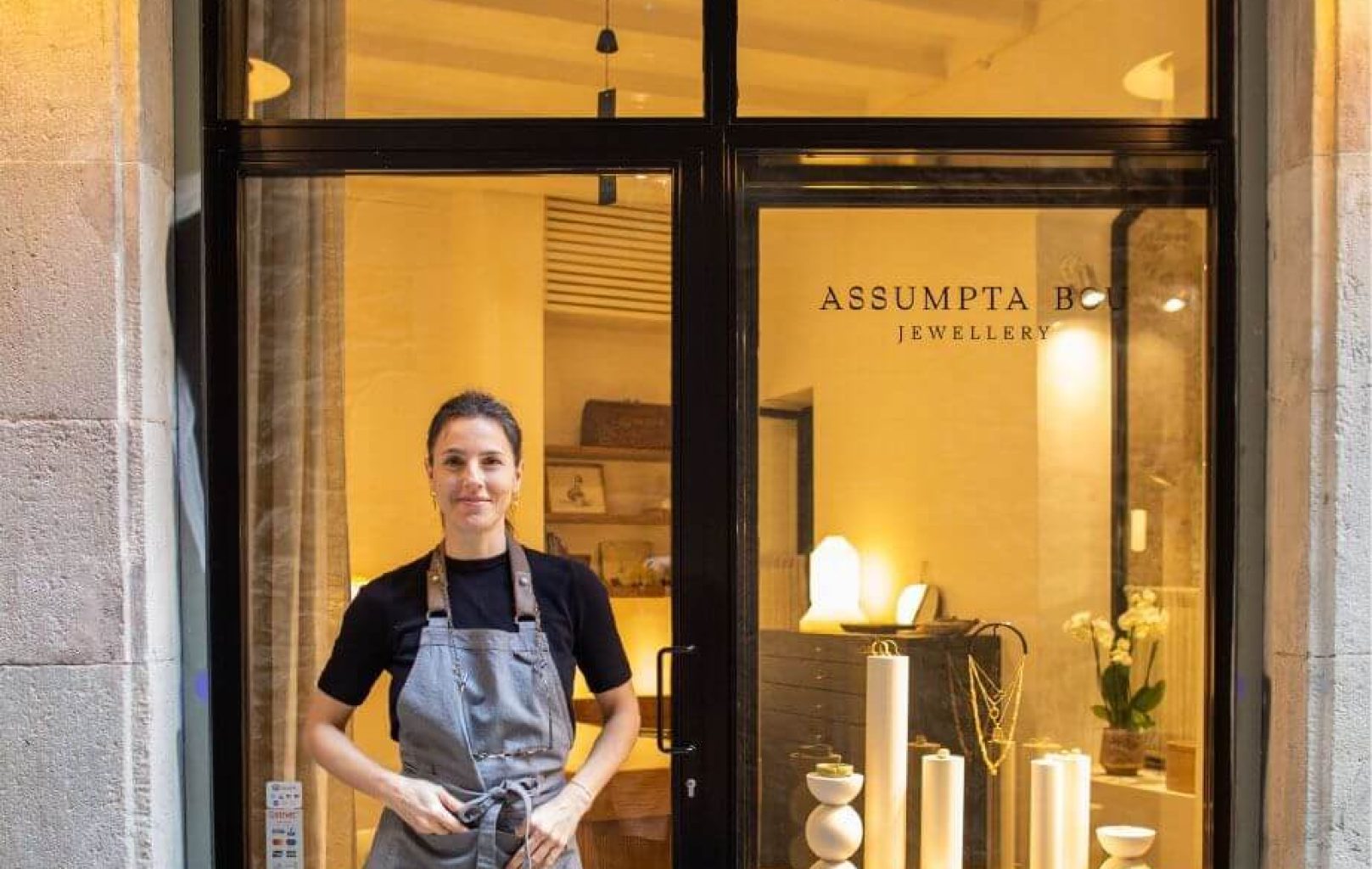A person wearing an apron stands in front of a jewelry shop with glass windows displaying decorative items and the sign "ASSUMPTA B JEWELLERY" by Assumpta Bou visible behind them.