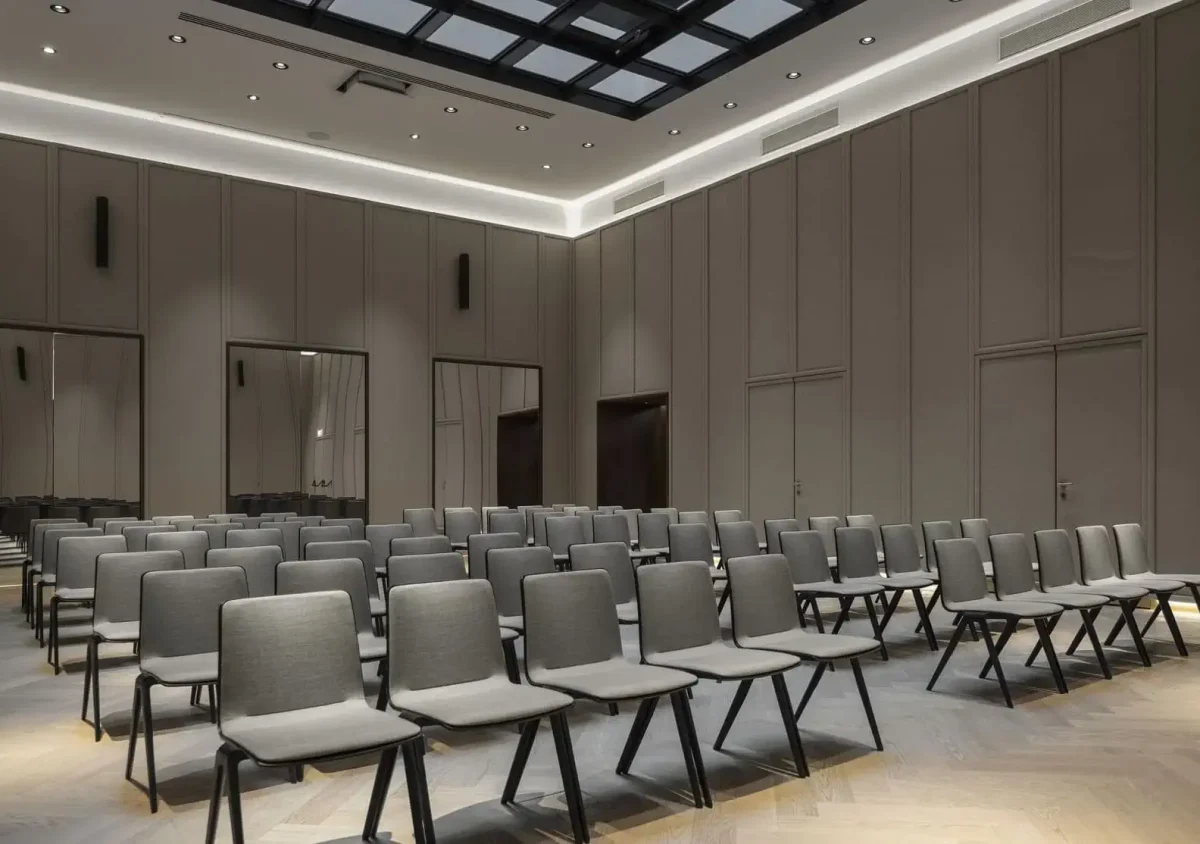 A modern conference room designed for events and meetings, featuring rows of empty gray chairs facing the front, high ceilings, and neutral-colored walls with recessed lighting.