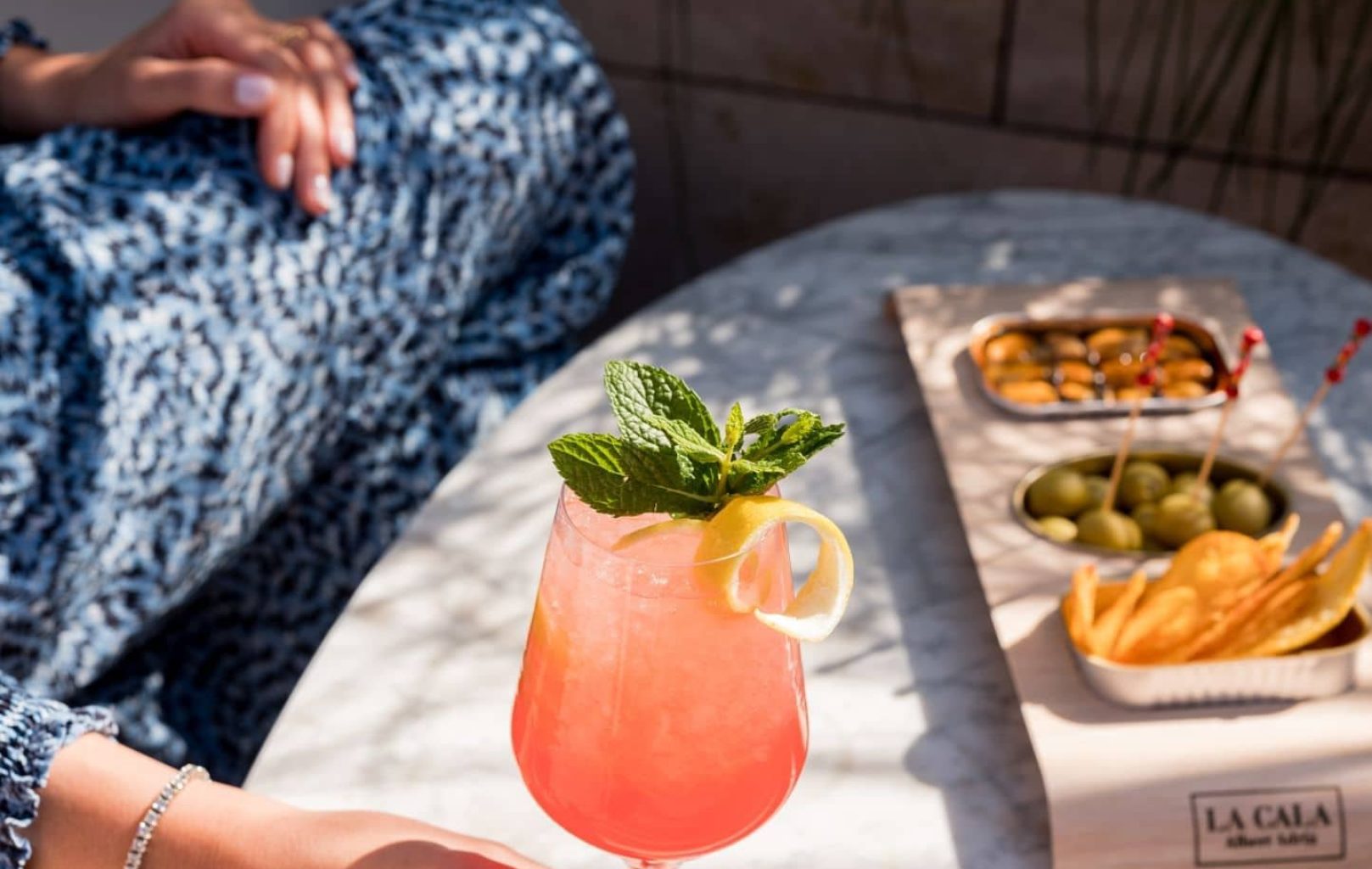 En el Azimuth Rooftop Bar, una persona sostiene un cóctel rosa adornado con menta y limón en una mesa de mármol, junto a una bandeja de patatas fritas, aceitunas y frutos secos.