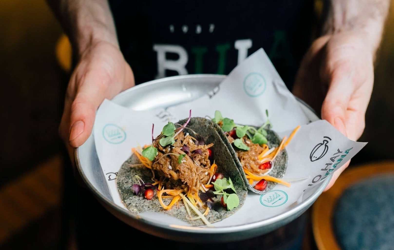 Una persona sostiene un plato con dos tacos rellenos de carne desmenuzada, microgreens y verduras sobre papel de la marca BABULA BAR 1937.