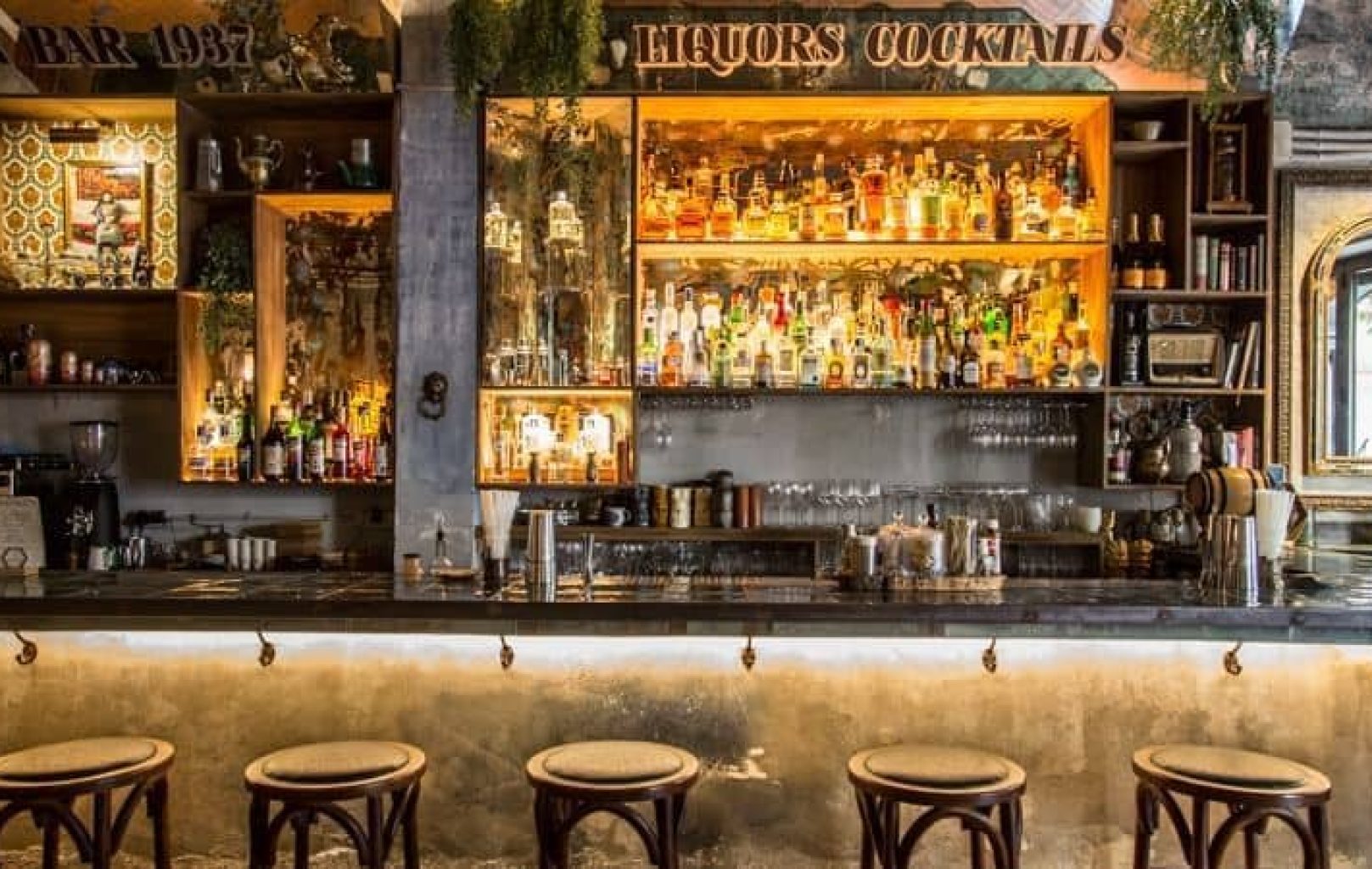 Un BAR 1937 bien iluminado con cinco taburetes, una encimera de mármol, estanterías repletas de botellas de licor y elementos de decoración vintage.