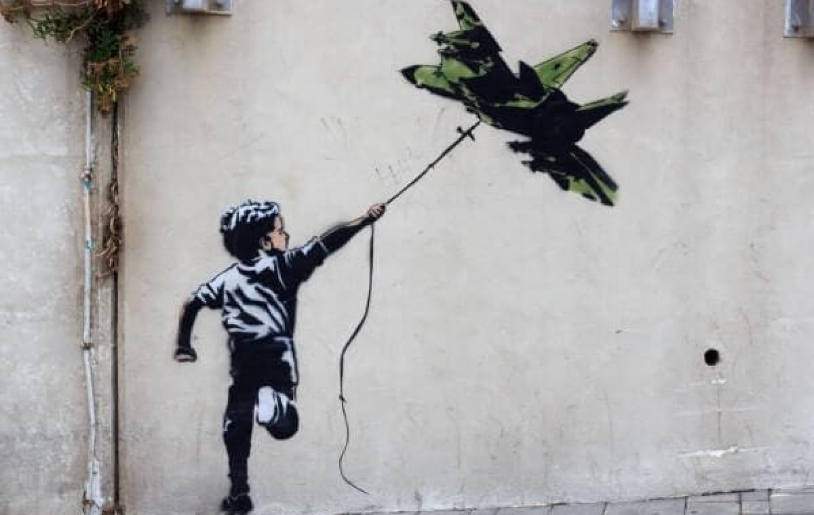 Street art inspired by Banksy: a child holds a string tied to a military fighter jet, floating like a balloon, symbolizing Art of Protest on a beige wall.