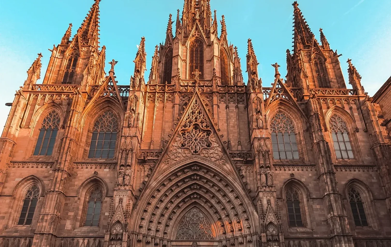 La gent es troba davant d'una gran catedral gòtica amb arcs apuntats, maçoneria intricada i altes agulles, sota un cel blau clar.