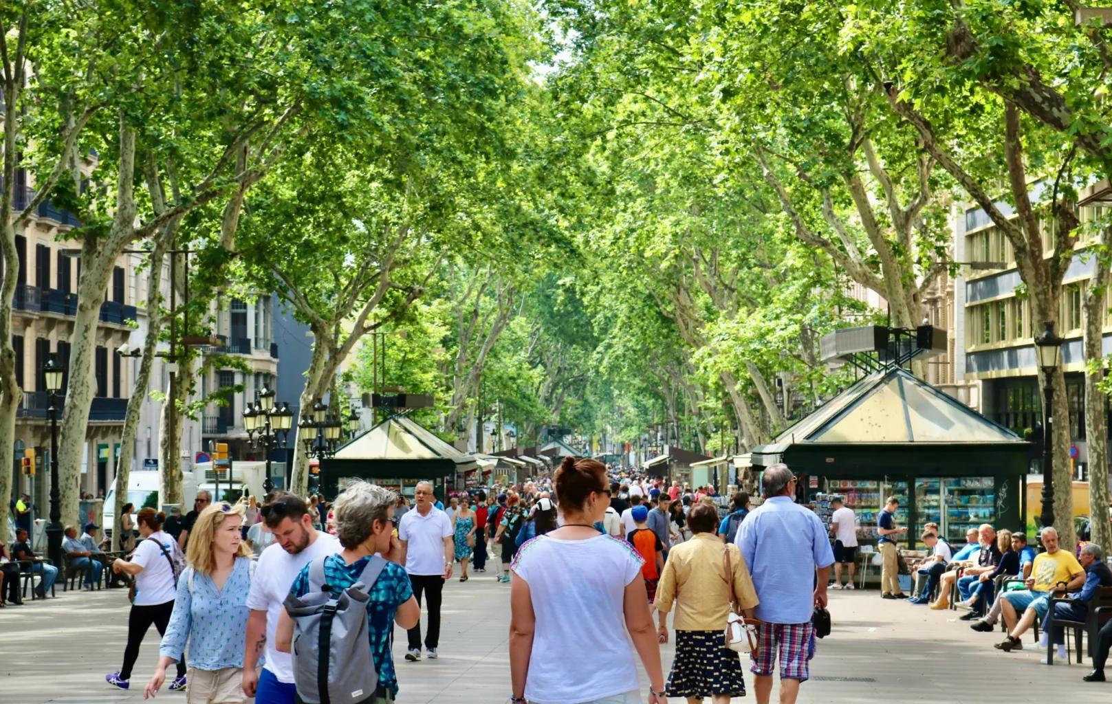 Multitut de gent caminen per un carrer de vianants amb arbres i quioscos verds i botigues a banda i banda durant el dia.