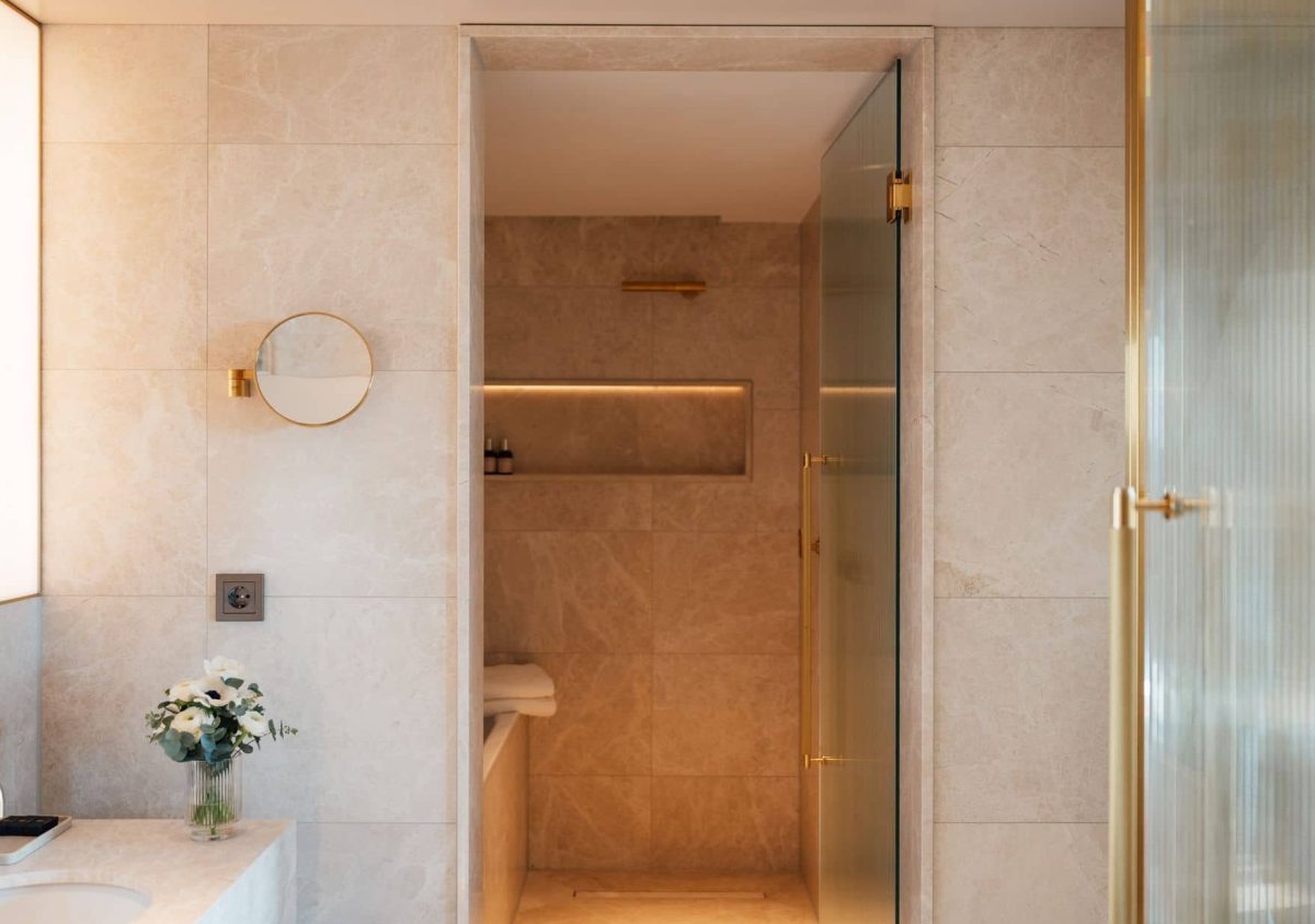 Modern bathroom in the Almanac Room with beige marble walls, a walk-in shower with glass door, a round mirror, and a small vase of flowers on the counter.