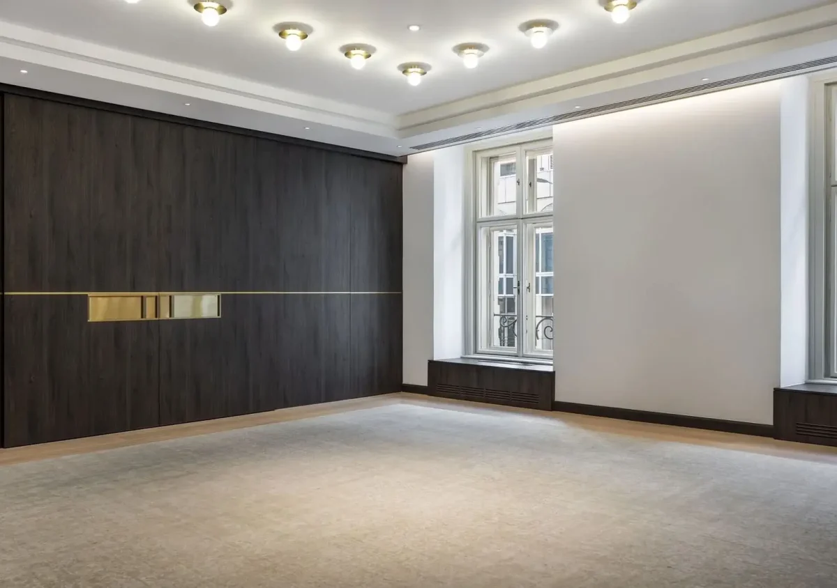 A modern, empty room with large windows, a beige carpet, dark wood paneling on one wall, and ceiling lights.
