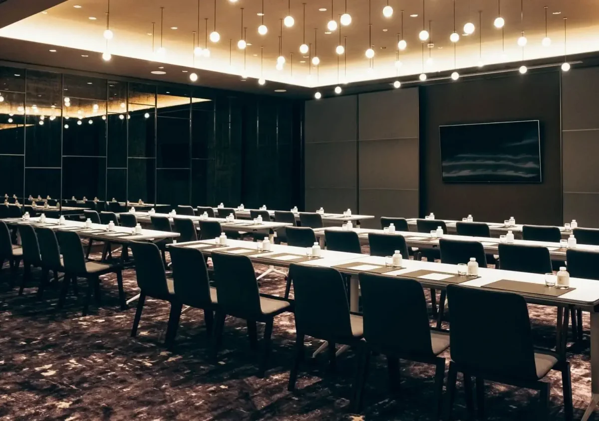 A modern conference room with a pre-function area, featuring rows of tables and chairs set with notepads and water bottles under ceiling lights.